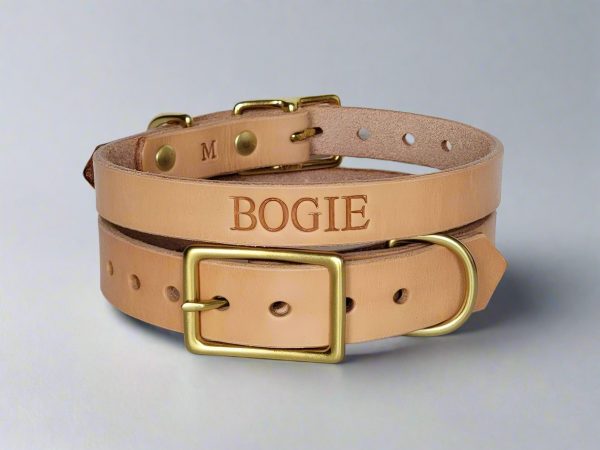 Handmade natural leather dog collar with solid brass hardware, personalized with the name "BOGIE" stamped into the leather. Collar is displayed coiled on a white background, showing slicked edges, brass buckle, and D-ring attachment.